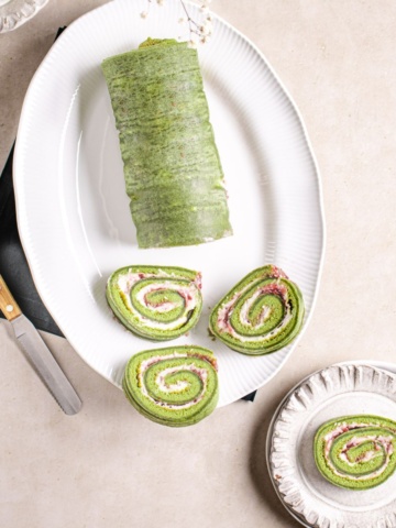 Matcha roll cake on a white platter with knife, and serving dishes.