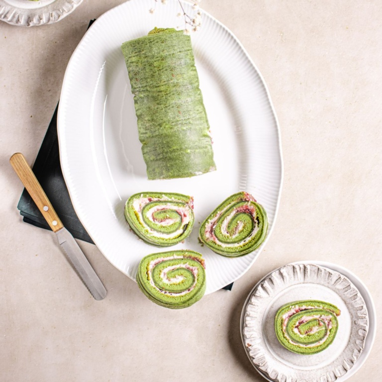 Matcha roll cake on a white platter with knife, and serving dishes.