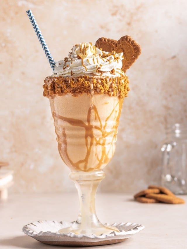A cup of lotus milkshake with whipped cream drizzled with cookie butter spread in a tall glass.