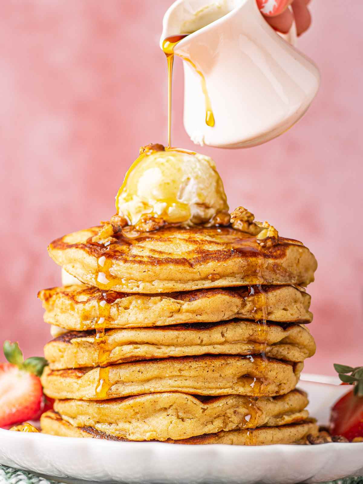 Stack of oat meal pancakes on plate with whipped topping while drizzling maple syrup.